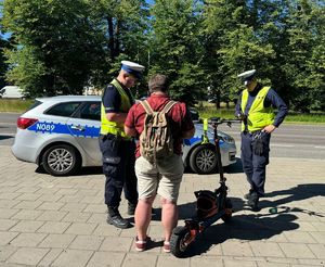 Policjanci ruchu drogowego kontrolują kierującego hulajnoga, w tle radiowóz